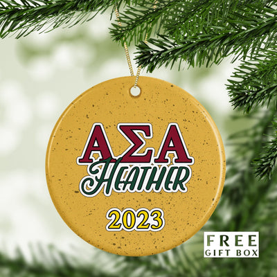 May include: A round, yellow Christmas ornament with a red, white, and green Greek letter design that reads &#39;AZA Heather 2023&#39;.
