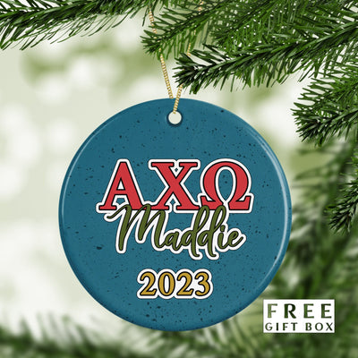 May include: A blue and white ceramic Christmas ornament with the letters AXQ in red and white, the word Maddie in green, and the year 2023 in gold. The ornament has a speckled blue background.