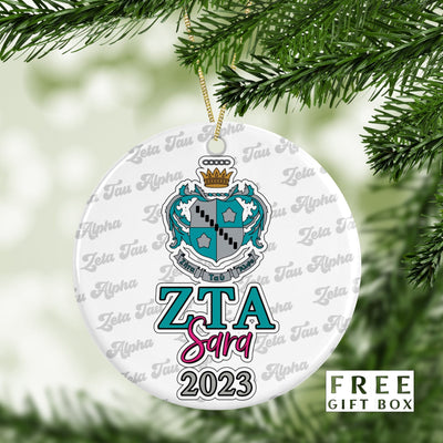 May include: A white round Christmas ornament with a teal and silver crest and the letters ZTA in teal with the word &#39;Sara&#39; in pink and the year 2023 in gray. The ornament has a repeating pattern of the words &#39;Zeta Tau Alpha&#39; in a gray font. The text &#39;FREE GIFT BOX&#39; is printed in black at the bottom of the ornament.