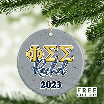 May include: A gray, round, ceramic Christmas ornament with a yellow and blue Greek letter design that reads &#39;ΦΣΣ&#39; and the name &#39;Rachel&#39; in blue script. The year &#39;2023&#39; is written in blue below the name.