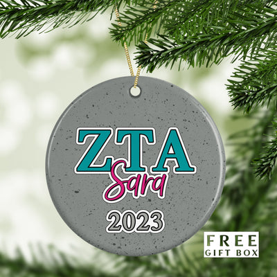 May include: A gray round ceramic ornament with a teal blue and white outline of the letters &#39;ZTA&#39; and the word &#39;Sara&#39; written in pink cursive underneath. The year &#39;2023&#39; is written in white underneath the word &#39;Sara&#39;. The ornament has a speckled gray background.