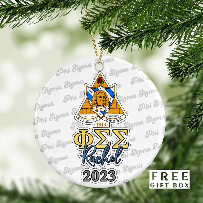 May include: A white round Christmas ornament with a gold and blue Phi Sigma Sigma sorority crest. The ornament has the letters &