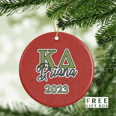 May include: A red round Christmas ornament with a green and white design. The ornament has the letters &#39;KA&#39; in green, the word &#39;Briana&#39; in black, and the year &#39;2023&#39; in white. The ornament is hanging from a gold string.