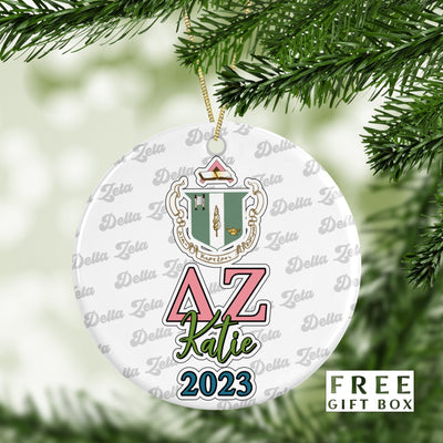 May include: A white round ceramic ornament with a green and pink sorority crest, the letters AZ in pink and green, the name Katie in green, and the year 2023 in blue. The ornament is surrounded by a repeating pattern of the words &#39;Delta Zeta&#39; in gray. The text &#39;FREE GIFT BOX&#39; is in the bottom right corner of the image.