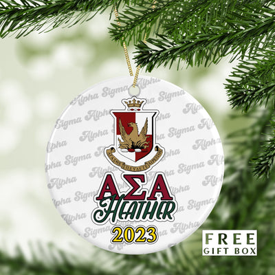 May include: A white round Christmas ornament with a red and gold crest, the letters &#39;ΑΣΑ&#39; in green, the name &#39;HEATHER&#39; in green, and the year &#39;2023&#39; in gold. The ornament is surrounded by a repeating pattern of the words &#39;Alpha Sigma Alpha&#39; in a white cursive font. The text &#39;FREE GIFT BOX&#39; is in the lower right corner of the image.
