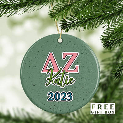 May include: A green round ceramic ornament with a pink and white Greek letter design that says &#39;AZ&#39; and the name &#39;Katie&#39; in green cursive writing. The year &#39;2023&#39; is written in blue and white below the name.