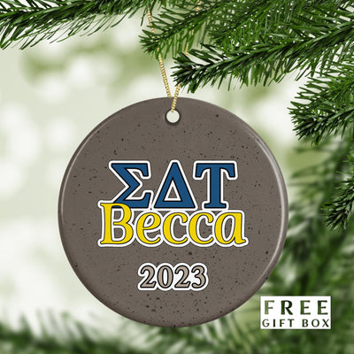 May include: A round gray ceramic ornament with a white string for hanging. The ornament has a blue and yellow design with the letters &#39;ΣΔΤ&#39; and the name &#39;Becca&#39; in yellow. The year &#39;2023&#39; is written in white below the name.