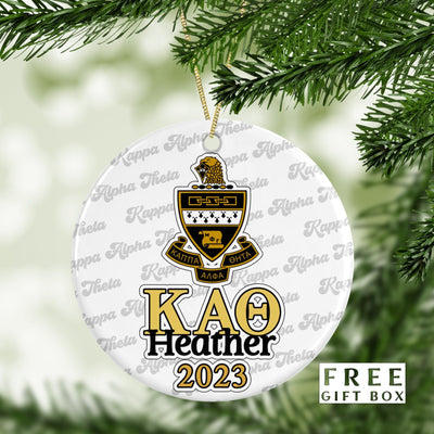 May include: A white round ornament with a gold and black Kappa Alpha Theta crest. The ornament has the letters &#39;KAΘ&#39; in gold with the name &#39;Heather&#39; and the year &#39;2023&#39; in black below. The ornament is surrounded by a repeating pattern of &#39;Kappa Alpha Theta&#39; in a cursive font. The ornament is hanging from a gold string on a green pine tree branch. The text &#39;FREE GIFT BOX&#39; is in the lower right corner of the image.
